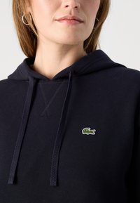 Sudadera azul marino de tela texturada, con un pequeño logotipo bordado de cocodrilo en verde y blanco en el pecho.
