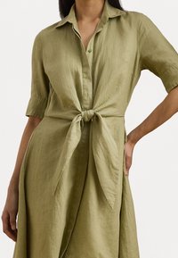 Abito camicia in lino verde oliva con nodo davanti, maniche corte, patta con bottoni e vestibilità morbida. Tessuto liscio e design semplice.