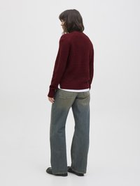 Suéter tricotado bordeaux com uma bainha ajustada, usado com jeans azuis desgastados e largos. Os jeans apresentam dois bolsos traseiros. Sapatos pretos complementam o look.