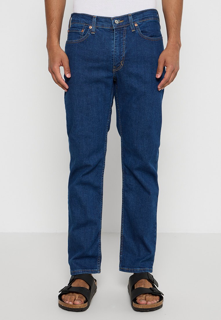 Dunkelblaue Jeans mit geradem Schnitt, kupferfarbenen Nähten und Knopfverschluss, kombiniert mit schwarzen Sandalen auf baren Füßen.
