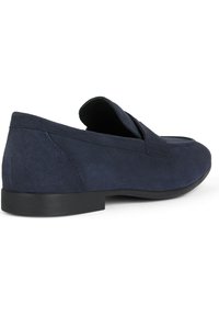 Scarpe slip-on in suede blu con una texture liscia, suola nera piatta e cuciture sottili sulla parte superiore. Design con cinturino aperto.