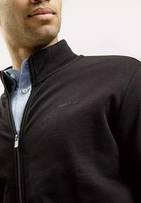 Czarna kurtka zapinana na zamek z wysokim kołnierzem, charakteryzująca się gładką fakturą. Dyskretnie wyszywane logo "CROSS JEANS" na piersi. Jasnoniebieska koszula pod spodem.