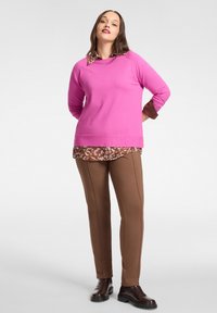 Różowy sweter z długimi rękawami nałożony na brązową koszulę w zwierzęcy wzór, zestawiony z dopasowanymi brązowymi spodniami i czarnymi botkami za kostkę.