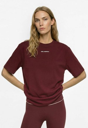 Kvinde med langt bølget hår iført en mørkerød Karl Lagerfeld T-shirt og matchende leggings, stående med hænderne i siden foran en hvid baggrund.