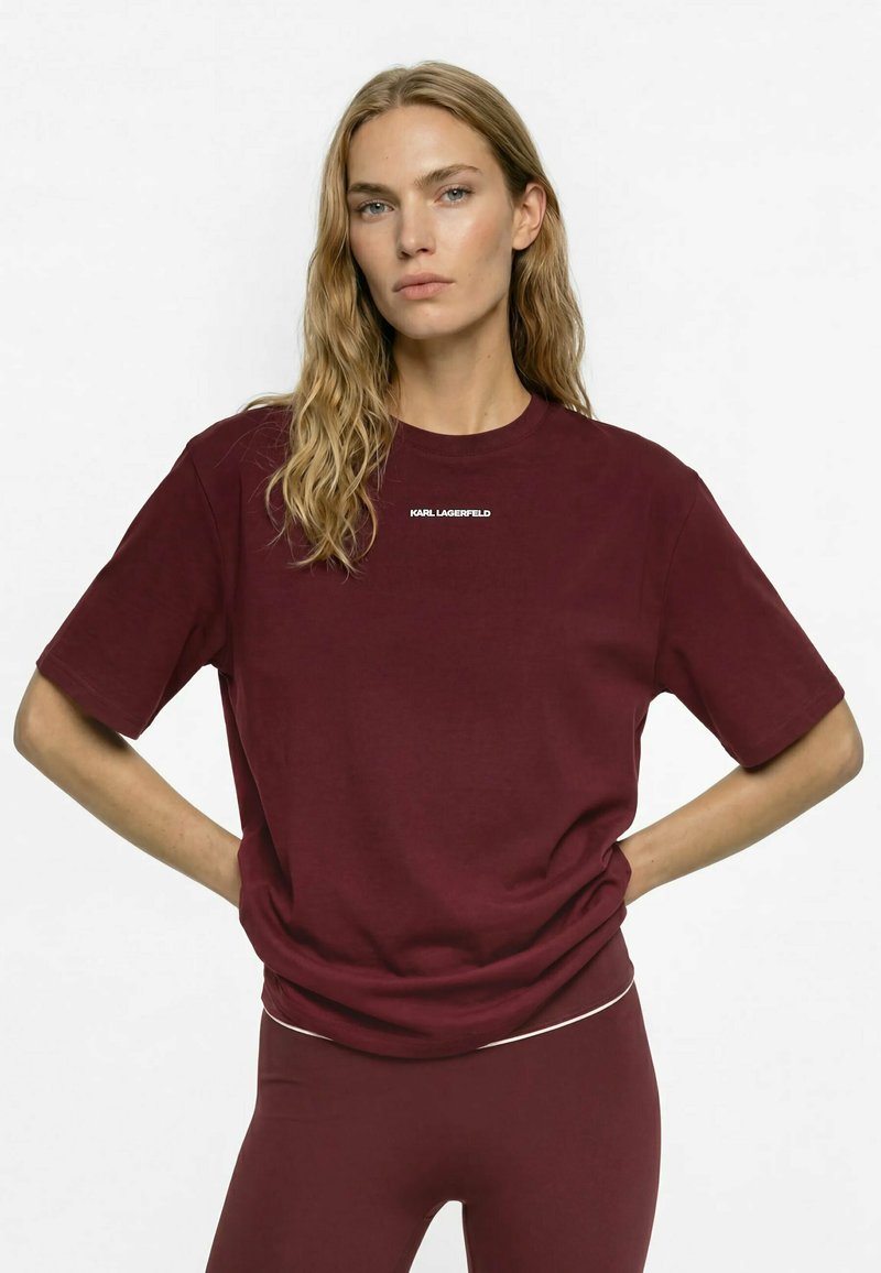 Femme aux longs cheveux ondulés portant un t-shirt bordeaux Karl Lagerfeld et des leggings assortis, debout les mains sur les hanches devant un fond blanc.