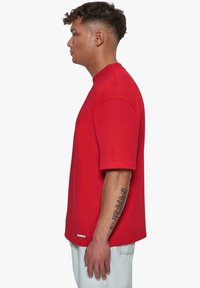 Camiseta de algodón roja con un corte relajado, mangas cortas y un pequeño logotipo en relieve en la manga. La textura parece suave y sin patrón.