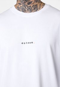Weißes Baumwoll-T-Shirt mit einem kleinen schwarzen und orangefarbenen gestickten Text, der "esteem" (Wertschätzung) liest. Rundhalsausschnitt und kurze Ärmel.