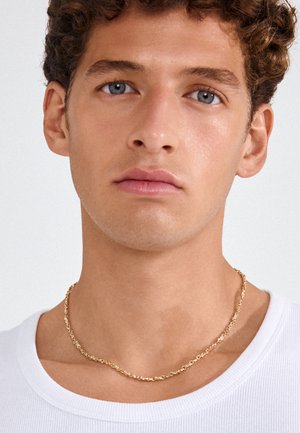 Mladý muž s kučeravými vlasmi a modrými očami, ktorý nosí zlatý reťazový náhrdelník a biele rebrované tričko na jednoliatom pozadí.
