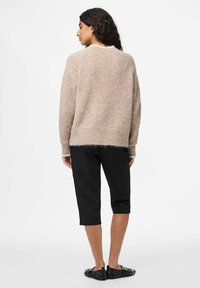 Ženska s temnimi lasmi nosi bež puhasto pulover, črne kratke hlače in črne ravne čevlje, stoji ob enobarvni beli steni.