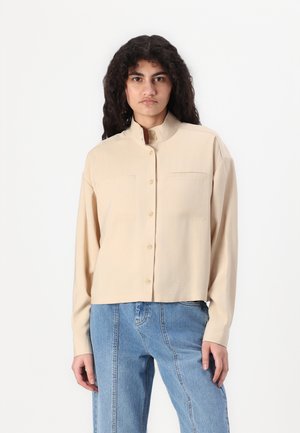 Beige long-sleeve shirt with a mandarin collar, featuring two chest pockets and button closure. Smooth fabric with a relaxed fit.