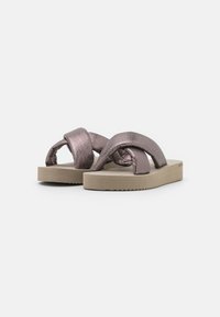 Sandales métalliques roses avec de larges sangles torsadées et une semelle beige minimaliste présentant un bord texturé. Un design plat et décontracté.