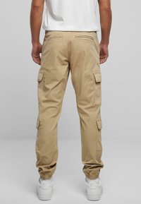 Khakifarbene Cargo-Hose mit elastischen Bündchen, mehreren Seitentaschen und einer lockeren Passform. Das Material wirkt strapazierfähig und leicht strukturiert. Weiße Sneakers sind sichtbar.