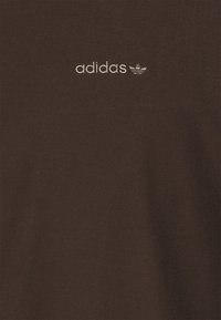 Brun bomullstyg med den broderade "adidas"-logotypen och trefoil-emblemet i krämfärg. Slät textur med subtil diagonal väv.