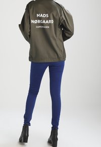 Veste bomber verte olive avec du texte blanc indiquant "MADS NØRGAARD COPENHAGUE" au dos, associée à un jean skinny bleu et des bottines noires.