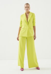 Limoengroene blazer met twee voorzakken, gecombineerd met bijpassende broeken met wijde pijpen. Model draagt roze stilettos. Effen witte achtergrond.