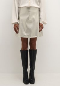 Fémes, világos bézs szoknya sima textúrával és pliszírozott szegéllyel, magas fekete fényes csizmával és világos, kötött, hosszú ujjú felsővel párosítva.