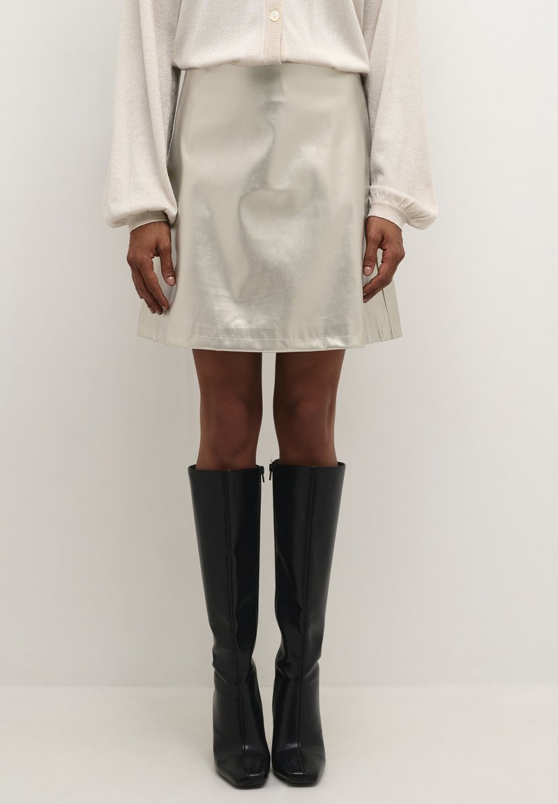 Fémes, világos bézs szoknya sima textúrával és pliszírozott szegéllyel, magas fekete fényes csizmával és világos, kötött, hosszú ujjú felsővel párosítva.