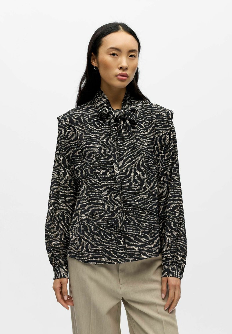 Mustakultainen kuvioitu pusero, jossa on rusetti kauluksessa, pitkät hihat ja väljä istuvuus. Kangas on teksturoitu ja aaltoileva.