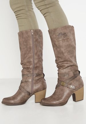 Stivali marroni testurizzati fino al ginocchio con cerniere laterali, cinturini decorativi con motivo a serpente, tacchi a blocco, indossati con pantaloni kaki chiari.