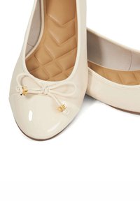 Beige patent leather ballet flats with a rounded toe, bow detail, and gold hardware accents. Textured beige insole with a zigzag pattern.