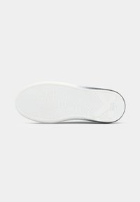 Λευκή σόλα αθλητικού παπουτσιού με υφή καουτσούκ, διαθέτοντας κυματιστό σχέδιο και γκρι λεπτομέρεια κοντά στη φτέρνα· φέρει τη μάρκα "Karl" κοντά στο πίσω μέρος.