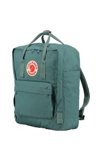Sac à dos en tissu vert de forme rectangulaire, avec une poche avant, des poignées supérieures et des bretelles réglables. Il présente un logo circulaire avec un fond rouge.