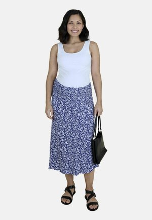 Uśmiechnięta kobieta w białym, bezrękawnym topie, niebieskiej spódnicy midi w kwiaty, czarnych sandałach, trzymająca czarną torebkę na jednolitym tle.