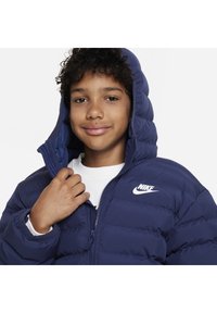 Giacca puffer blu con cappuccio, caratterizzata da un logo Nike bianco sul petto e una finitura trapuntata e testurizzata. Chiusura con zip, vestibilità aderente.