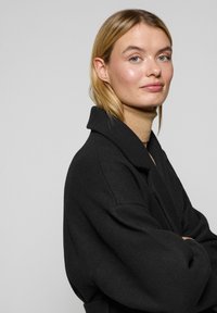 Abrigo de mezcla de lana negro con cuello estructurado, mangas holgadas y un acabado texturizado, llevado sobre un suéter de cuello alto beige.