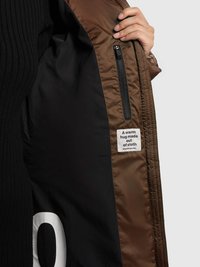 Intérieur d'une veste matelassée marron, avec une doublure noire lisse, une poche zippée et une étiquette en tissu portant le texte "Un câlin chaleureux fait de tissu."