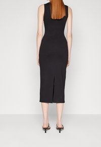 Svart, ärmlös midiklänning med en figurnära silhuett, med en bakre slits för rörelse. Slät tyg med minimal detaljering.