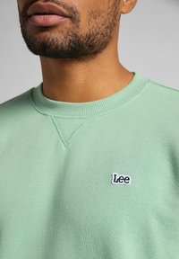 Sudadera verde menta con cuello redondo y detalles acanalados, que presenta un pequeño logo en blanco y negro de "Lee" en la parte frontal.
