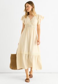 Beige maxi dress with flutter sleeves, lace detailing at the waist, flared hem. Worn with brown slides and a woven bag. Smooth fabric texture.