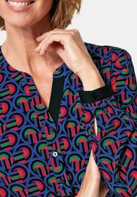 Kleurrijke blouse met een geometrisch patroon in rood, groen en blauw op een donkere achtergrond. Zwarte kraag en manchetten met knoopdetail.