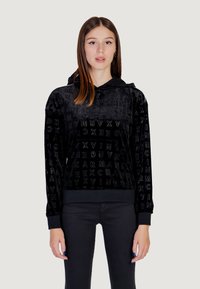 Sudadera de terciopelo negro con patrón de logo en relieve. Cuenta con capucha, mangas largas y puños y dobladillo acanalados. Combinada con pantalones negros.