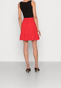 Rode uitlopende rok met ruches aan de zoom, gecombineerd met een nauwsluitend zwart tanktopje. Het model staat in zwarte mules met hak, waarbij de achterkant wordt getoond.