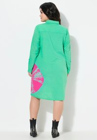 Robe chemise verte avec un motif circulaire rose sur le côté, à manches longues et poignets boutonnés. Portée avec des bottines noires.