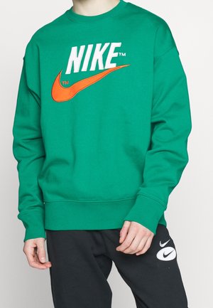 Osoba ubrana w zieloną bluzę Nike z dużym pomarańczowym logo Swoosh oraz czarne spodnie z białymi naszywkami Nike Swoosh na udzie.