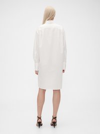 Abito camicia in cotone bianco con colletto, maniche lunghe e taglio dritto, abbinato a sandali neri con tacco.