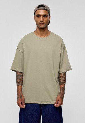 Khaki oversized t-shirt, van katoen, ronde halslijn, korte mouwen. Model draagt donkere denim shorts, zichtbare arm tattoos. Neutrale achtergrond.