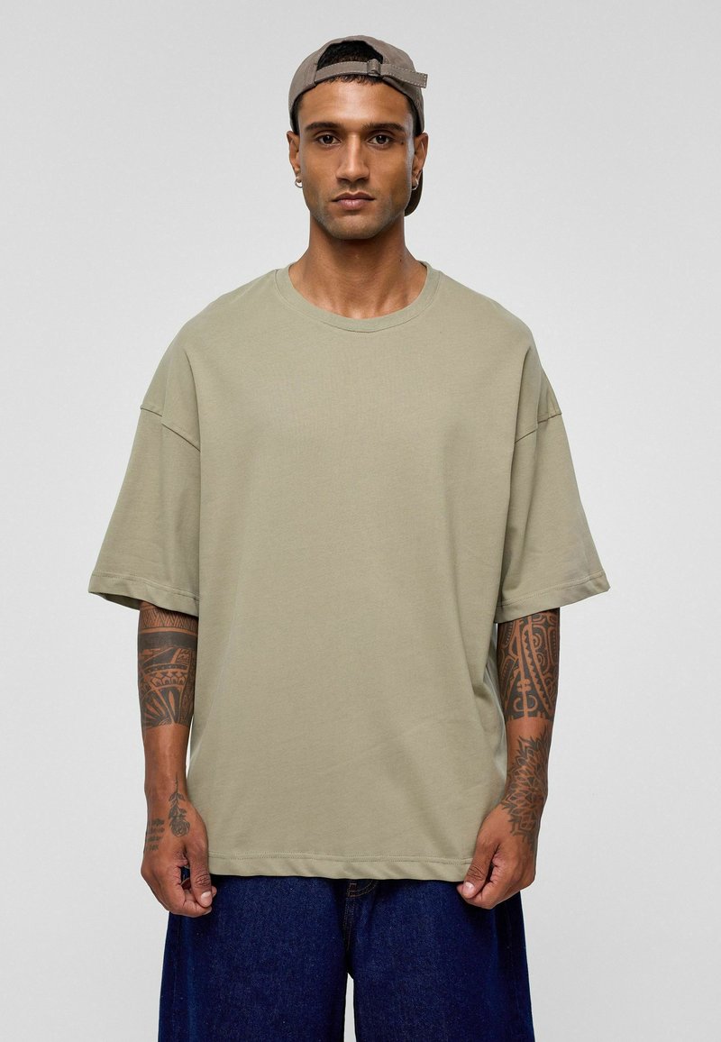Khaki oversized t-shirt, van katoen, ronde halslijn, korte mouwen. Model draagt donkere denim shorts, zichtbare arm tattoos. Neutrale achtergrond.