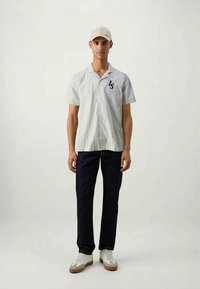 Camisa abotoada com padrão riscado claro, mangas curtas e logotipo preto. Jeans escuros e ténis brancos completam o visual. Boné simples.