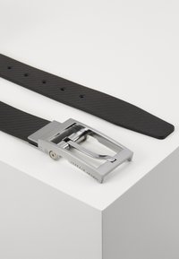 Zwarte rubberen riem met een gestructureerd oppervlak, voorzien van een zilveren metalen gesp en een strak ontwerp, geplaatst op een witte ondergrond.