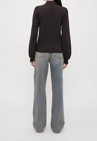 Frau in einem schwarzen Langarmoberteil und hochgeschnittenen, hellblauen weit geschnittenen Jeans, die mit dem Rücken zu einer schlichten Hintergrund steht.