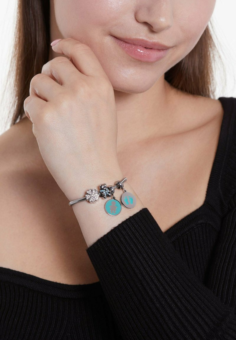 Silberner Charm-Armband mit einer Blume, verschiedenen Perlen und zwei türkisfarbenen runden Charms mit Symbolen. Getragen an einem Handgelenk mit einem schwarzen Pullover.