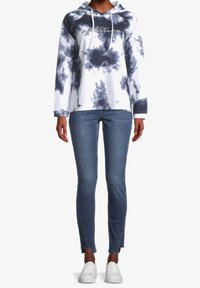 Sudadera blanca con capucha y patrón de tie-dye azul marino, con texto que dice "ambición". Combinada con jeans ajustados azules y zapatillas blancas.