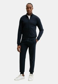 Uomo in piedi che indossa una giacca a zip blu navy, pantaloni coordinati, una maglietta bianca sotto e sneakers bianche, con una mano in tasca.