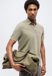 Νεαρός άνδρας με κοντό αφρο, φορώντας μπεζ πόλο πουκάμισο, κρατά μια μεγάλη δερμάτινη τσάντα ταξιδίου σε ελιά πράσινη και καφέ από τα χερούλια της.