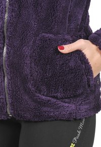 Veste en polaire violette avec une surface texturée et pelucheuse. Elle dispose d'une fermeture éclair sur le devant et de deux poches latérales, mettant en valeur des coutures centrées.