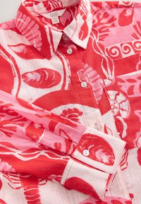 Camicia in lino a fantasia rossa e rosa con motivi di conchiglie e coralli, collo appuntito e bottoni bianchi sul davanti e sui polsini.